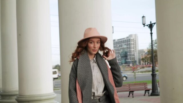 Portrait of a young woman with makeup in a hat and coat in the city against the background of tall columns. Slow mo