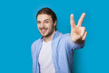 Fototapeta premium Handsome caucasian man with long hair and beard is gesturing the peace sign smiling at camera on a blue studio wall