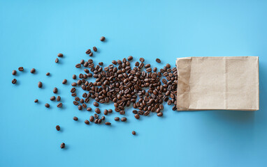 Flat lay of roasted coffee beans spilling from brown paper bag on blue background 