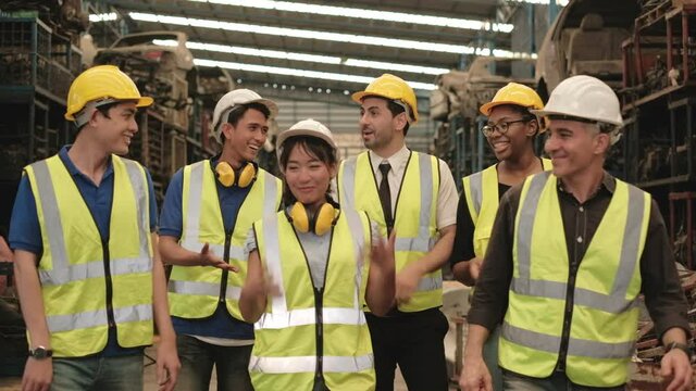Various races workers and engineers of factory are having fun joking around in machine-spare parts warehouse during break. The entire team wears safety uniforms and industrial helmets.