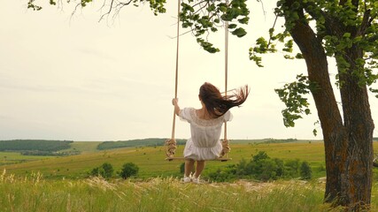 A young girl in dress swinging on swing in evening park. Wooden swing with swinging free, happy woman outdoors. Swing on a swing, dreams of flying. Travel in spring summer in nature. Healthy lifestyle - obrazy, fototapety, plakaty