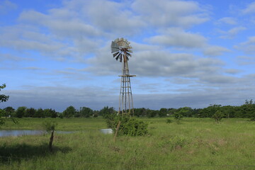 A Kansas Farm Windmill in a pasture with green grass, fence, tree,  with blue sky and clouds north of Hutchinson Kansas USA out in the country.