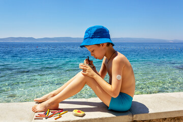 Diabetes.  A boy with sugar monitoring system sitting on the beach and draws on pebbles. Concept life with diabetes, glycemic control. Children's creativity in the fresh air on vacation © KRISTINA KUPTSEVICH
