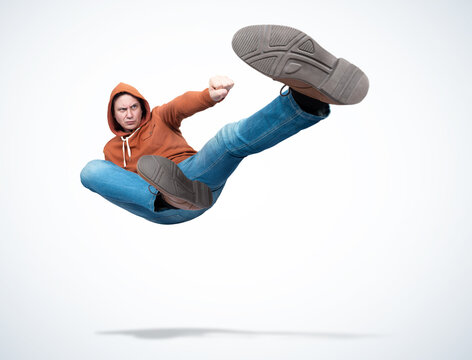 Man in orange hooded sweatshirt and jeans in karate fight jump, on light background. 