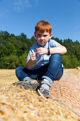 Naklejka na ściany i meble red-haired boy sitting on a golden straw stack in a field