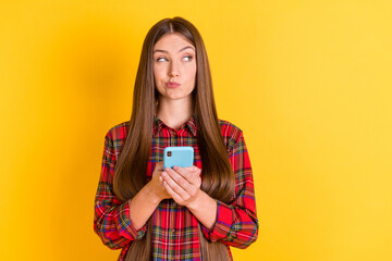Fototapeta premium Portrait of attractive skeptic minded girl wearing checked shirt using device deciding copy space isolated over bright yellow color background
