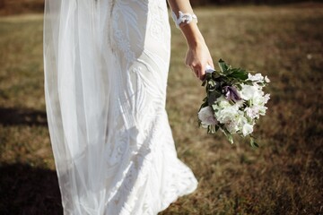 The bride girl in a white wedding long dress walks across the field and carries a bouquet of wildflowers in her right hand. No face, close up