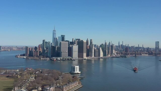 An aerial view of New York harbor on a sunny day with clear blue skies. The drone camera dolly in over the water, high enough to see Governors Island ventilation tower and Manhattan in the back.