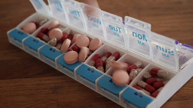 Multivitamin and prescription pills falling on weekly pill container in slow motion. Multivitamins and Creon digestive enzymes for Cystic Fibrosis daily treatment.