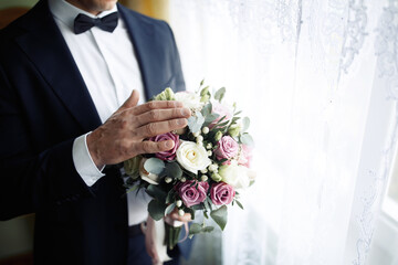 Man groom in a beautiful stylish classic suit holds a wedding bouquet of white and pink roses standing at the window waiting for the bride. Hands without a face.
