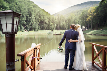 The groom and the pregnant bride in a white dress and blue suit hugging each other stand on the pier of a small lake and look into the distance at the mountains and sunset. 