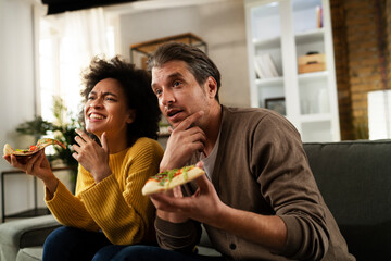 Cheerful young couple sitting on sofa at home. Happy woman and man eating pizza while watching a movie.