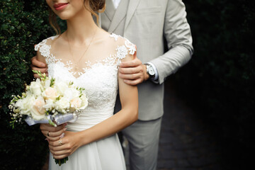 The bride and groom are standing next to each other and a man in a grey stylish suit with a bow-tie gently holds her by the shoulders. Girl in a white dress with a bouquet of roses flowers in her hand