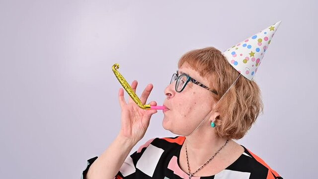 Happy elderly woman in a birthday cap blowing a whistle on a white background