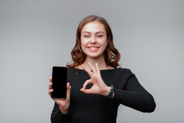 Portrait of a nice young brunette woman holding smartphone and showing OK sign, girl recommend upload new app