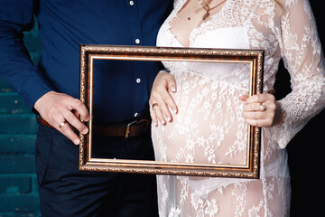 A man in a blue denim shirt and a woman in a white transparent peignoir hands make a heart on her pregnant belly and hold an empty golden picture frame on a background of a blue brick wall close-up
