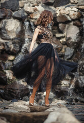 A girl with long slim legs in a shiny translucent black dress with a skirt and high-heeled shoes is dancing against the background of a waterfall and a stone wall.