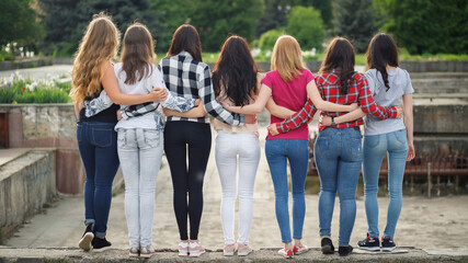 Seven athletic volleyball girls with beautiful priests of fit young slim girls in shirts and jeans hugging each other look into the distance with their backs to the camera.  
