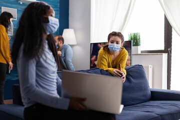 Happy cheerful woman holding beer bottle looking at laptop talking with african friend in apartment living room wearing face mask to prevent coronavirus spread in time of global pandemic. Conceptual