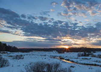 Bright rays of the sun on the horizon. Sunset, the sun shines through the clouds. Snow and bushes in the foreground. Early spring. Sunlight is reflected in the river.