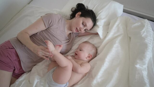 Happy young mother lying with cute infant toddler baby boy on bed, holding him on arms, hugging and playing early in morning. Healthy child, concept of hospital and happy motherhood. Nursery.