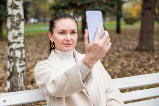 beautiful woman sits on a bench in the park. middle aged woman taking a selfie on the phone in the park