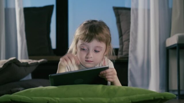 Curious Little Kid Girl Using Digital Pad Entertainment Device Lying On Sofa Alone. Small Child Hold Pad Computer Playing Games At Home. Children Tech Addiction Concept