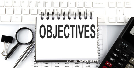 White notebook with text OBJECTIVES on the white keyboard with calculator,magnifier and pen