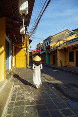 A young girl in traditional dress Ao Dai and conical hat standing on Hoi An ancient street