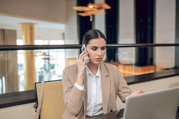 Young businesswoman in beige jacket talking on the phone and looking serious