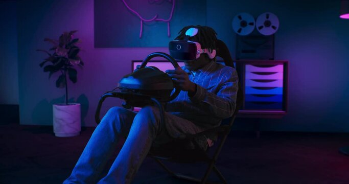 A guy is playing racing game in VR. He is sitting on the gaming chair with a steering wheel and pedals. A guy wears VR headset. Chair is located in the living room with red and blue neon lighting.
