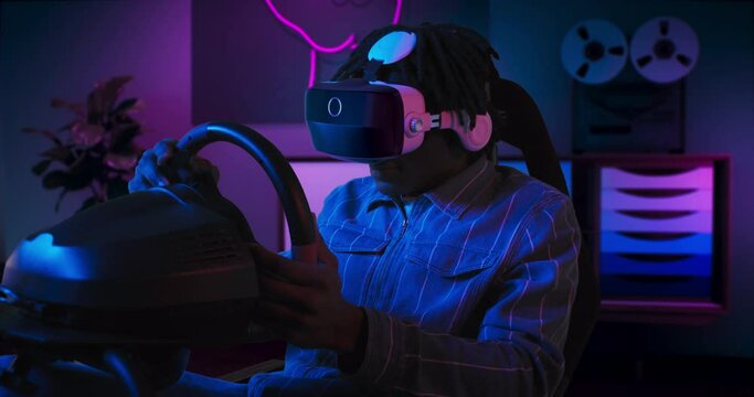 A guy is playing racing game in VR. He is sitting on the gaming chair with a steering wheel and pedals. A guy wears VR headset. Chair is located in the living room with red and blue neon lighting.
