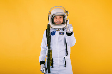 Male cosmonaut in spacesuit and helmet, raising thumb, on yellow background.