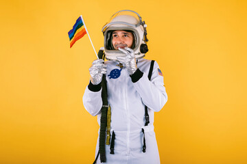 Gay male cosmonaut in space suit and helmet, holding lgtbi rainbow flag, on yellow background.