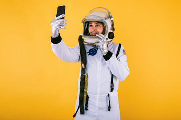 Male cosmonaut in space suit and helmet, taking a selfie with mobile phone, on yellow background. © Davidbenito