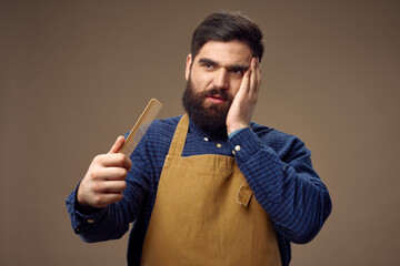 a man with a beard touches his face with his hand and a comb barbershop beige background apron