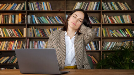 Concentrated woman freelancer types on grey laptop and stretches neck sitting on chair against large wooden racks with coloured books