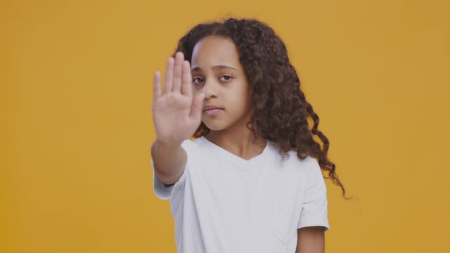 Serious african american curly girl gesturing STOP to camera and hsaking head in rejection, orange studio background