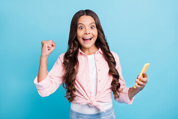 Photo of young excited crazy positive girl raise fist in victory with open mouth hold phone isolated on blue color background