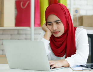 Portrait shot of young Muslim woman wearing a red hijab sitting on a black office chair, looking at the laptop with serious thinking about how to solve her company problems. Concept of stress at work