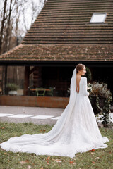 Obraz premium bride in a long white dress is standing in the park and holding a wedding bouquet of roses flowers and greens. young woman with hairstyle, makeup and long veil outdoors near hotel