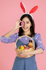 Young Asian woman wearing a Bunny hairband, holding an Easter egg, covering her eyes and hold a basket full of colorful eggs with a smiling face. The concept easter eggs festival with pink background