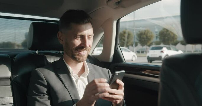 Close Up of a Relaxed Calm Businessman is texting Messages while traveling in the Car. A handsome Caucasian Businessman is using Smartphone in his Elegant Car while riding with his own Driver.