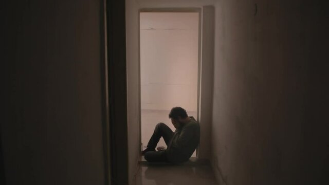 A sad desperate man, sitting on the floor near a door in his house, crying alone, letting his bad feelings come out.
