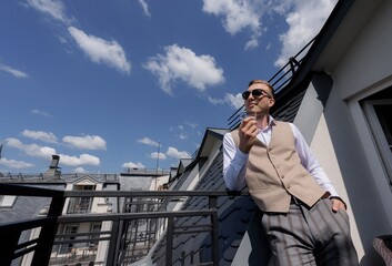 Front view of handsome man dressed in retro style costume standing on the balcony and looking at the city on the sky background.
