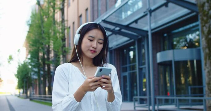 Attractive young woman in headphones using smartphone and listening to music. Joyful Asian lada tapping, browsing scrolling on Internet and shaking head to beats during city walk. Free time concept.