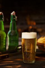 A tall glass of golden beer with a foam head on a table with empty green beer bottles and a bowl of chips in the background against a fire backdrop.