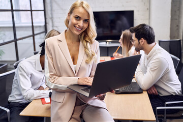 Confident young woman in formal clothes, working on laptop and looking on the camera sitting at his workplace in the office
