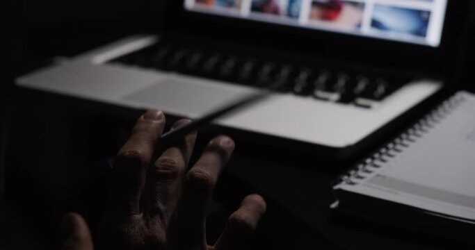 Dark lit footage of man hang spinning pen while working in from of laptop computer at late night. 