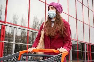 Young woman in medical mask and with shopping cart near supermarket. Concept of coronavirus epidemic